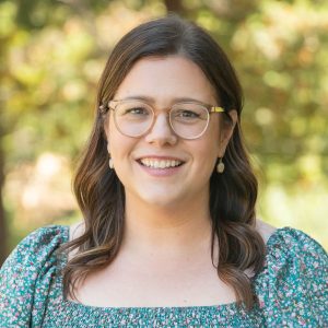 Smiling woman wearing clear-framed glasses outdoors, in a teal floral top, with a sunlit green background.