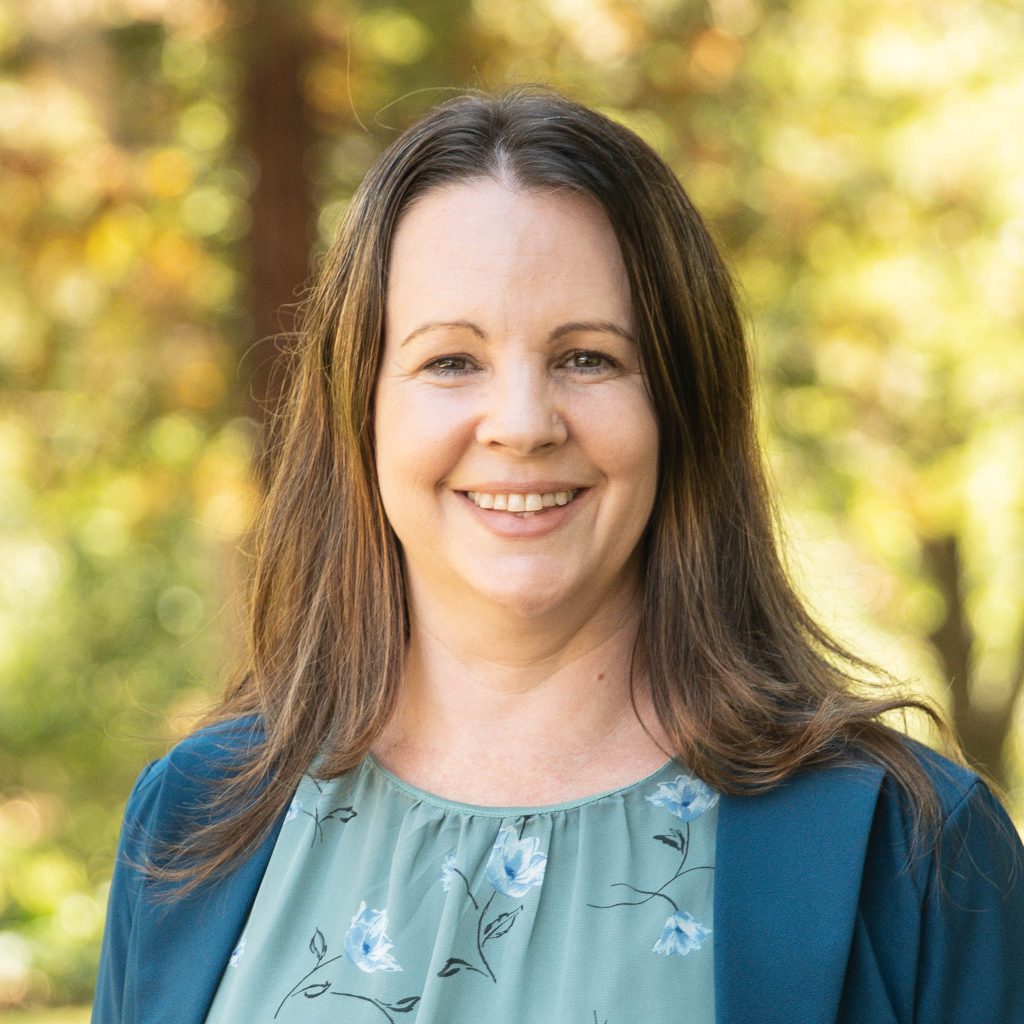 WendyR - Venture Christian Academy Smiling woman outdoors with autumn colored leaves in the background; close-up of her face and long brown hair, wearing a blue cardigan and teal blouse with floral pattern.