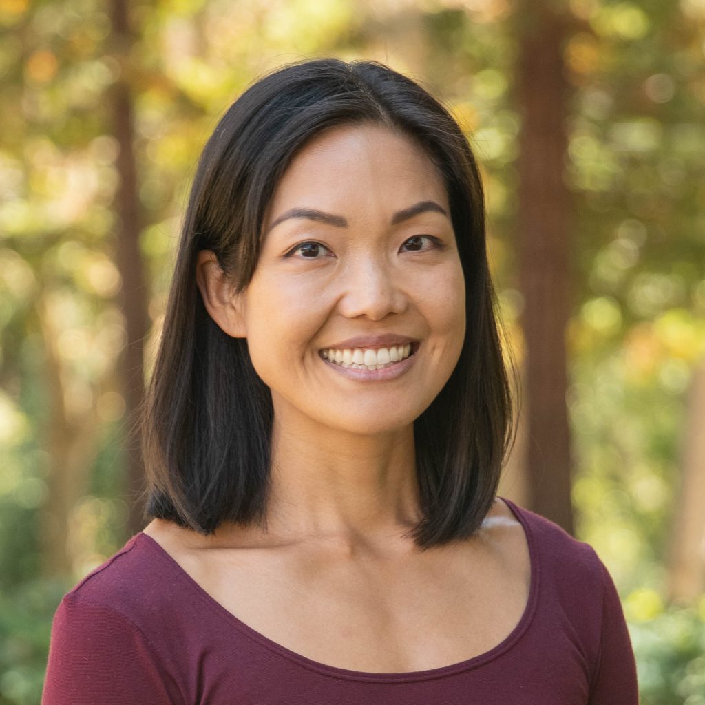 MichelleK - Venture Christian Academy Smiling woman outdoors wearing a burgundy top, with a blurred green and yellow leafy background.