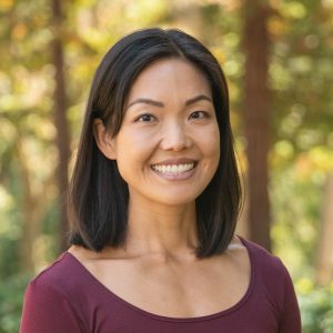 Smiling woman outdoors wearing a burgundy top, with a blurred green and yellow leafy background.